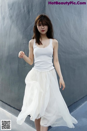 A woman leaning against a wall in a white tank top and skirt.