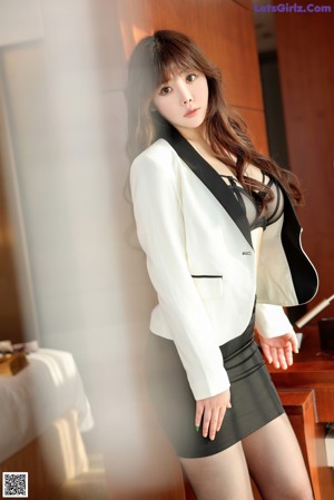 A woman in a white blazer and black pantyhose posing in a hotel room.