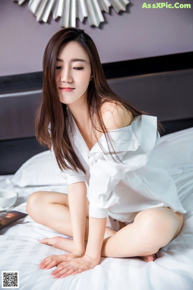 A woman sitting on a bed in a white shirt.